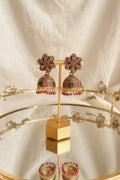 A pair of ornate gold-toned Indian jhumka earrings with pink, green, and black stone embellishments, displayed on a gold stand on a reflective glass table.