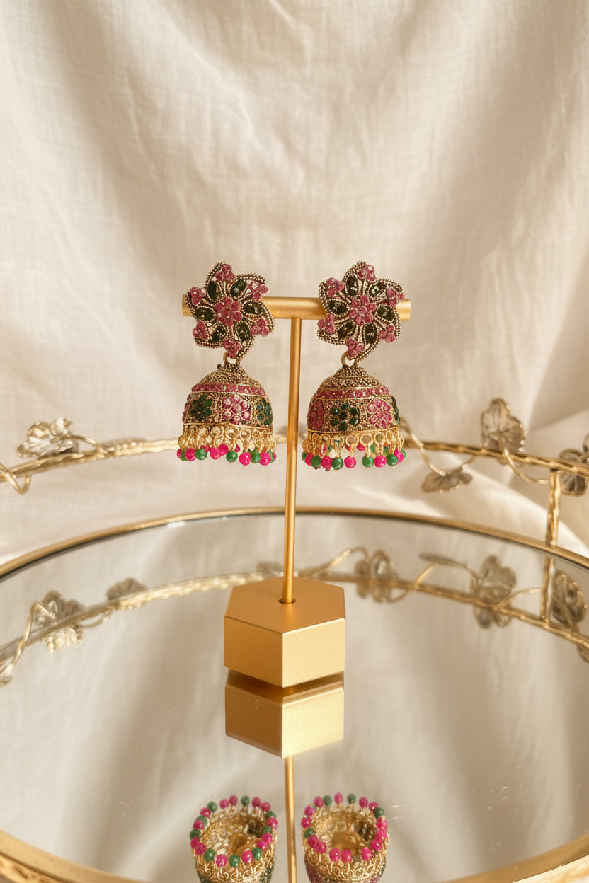 A pair of ornate gold-toned Indian jhumka earrings with pink, green, and black stone embellishments, displayed on a gold stand on a reflective glass table.