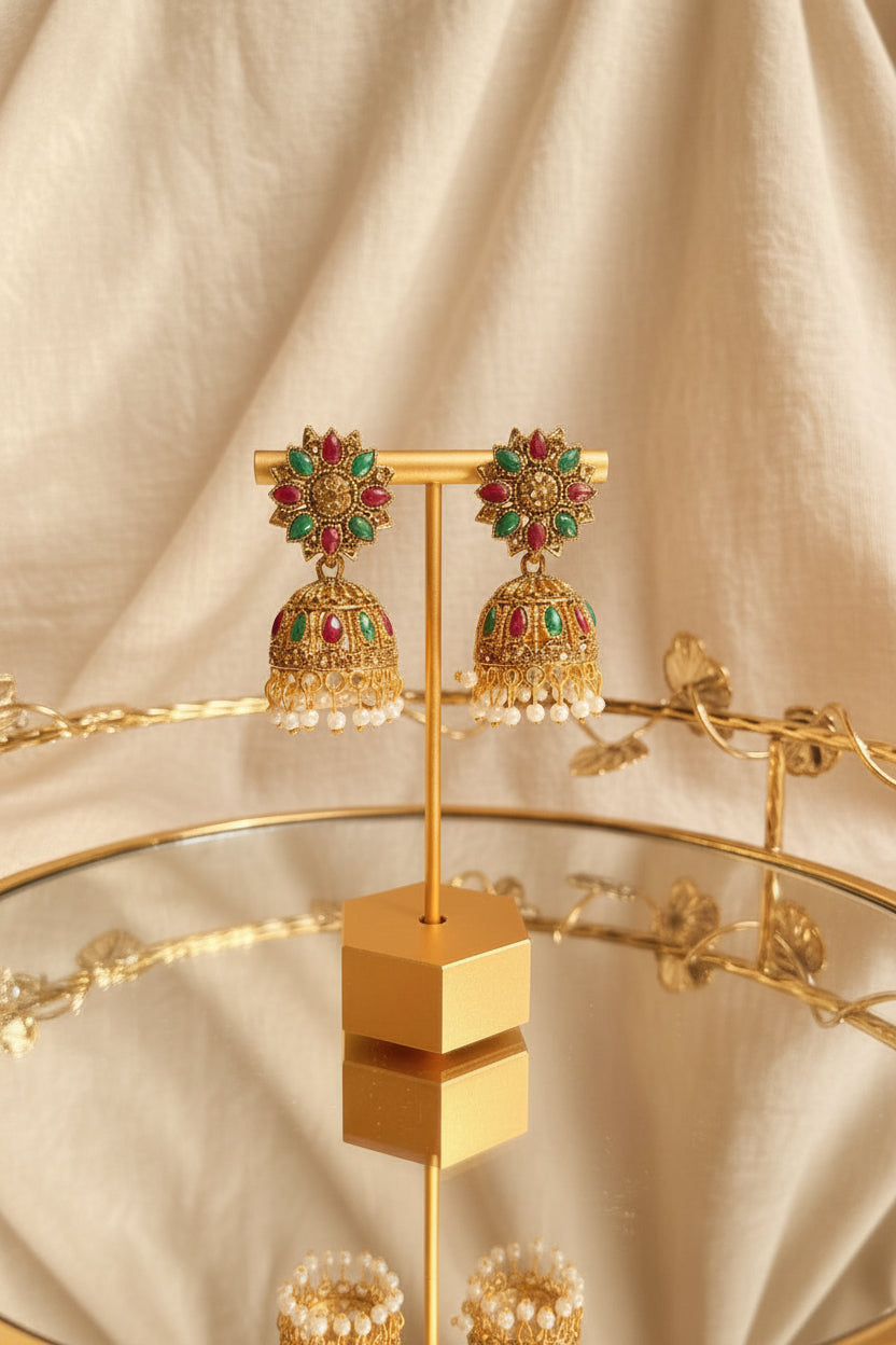 A pair of ornate gold-toned Indian jhumka earrings with red and green stones and white pearl drops, displayed on a gold stand on a reflective glass table.