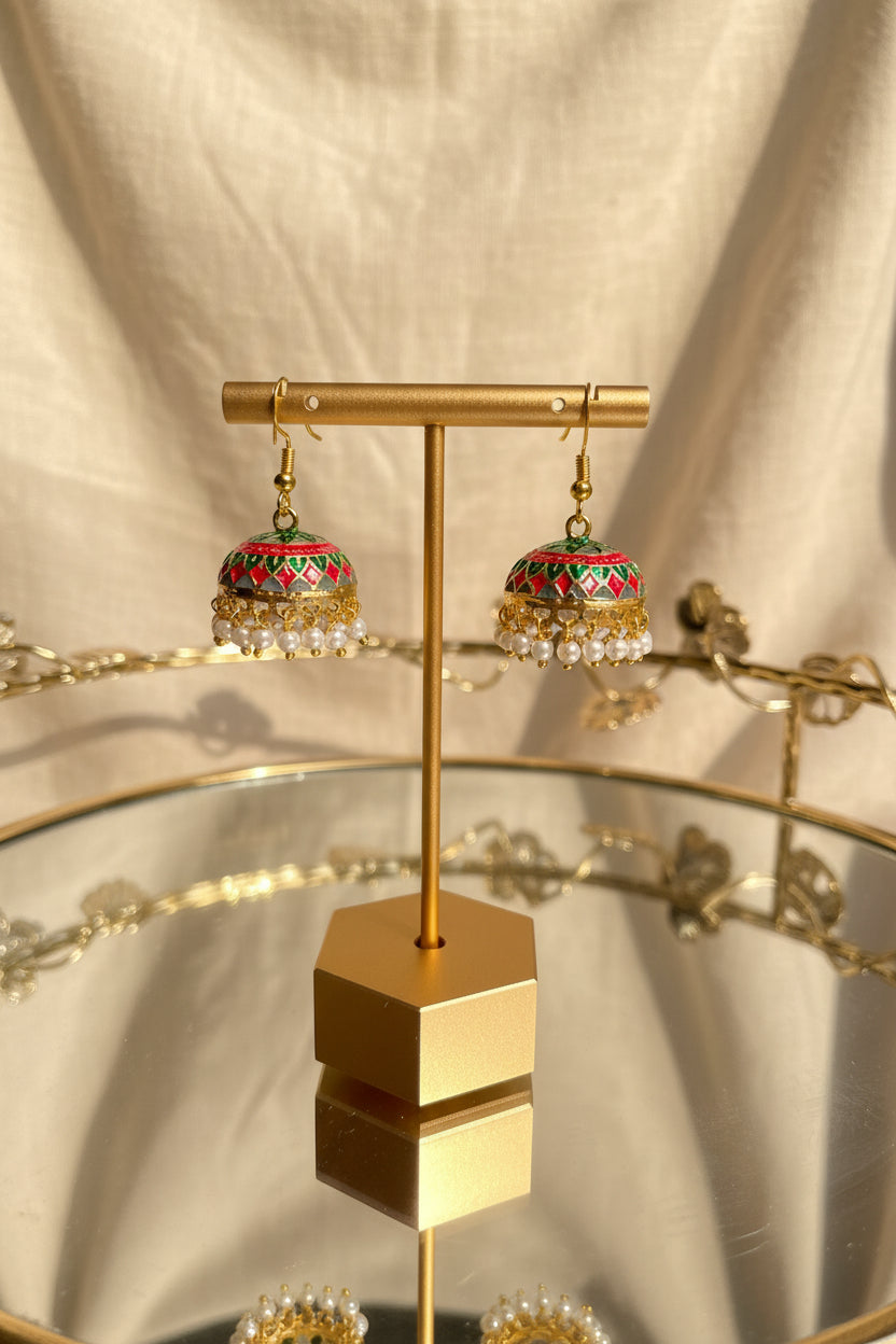 A pair of ornate gold-toned Jaipuri jhumkha earrings with red, green, and blue enamel detailing and white bead fringe, displayed on a gold stand on a reflective glass table.