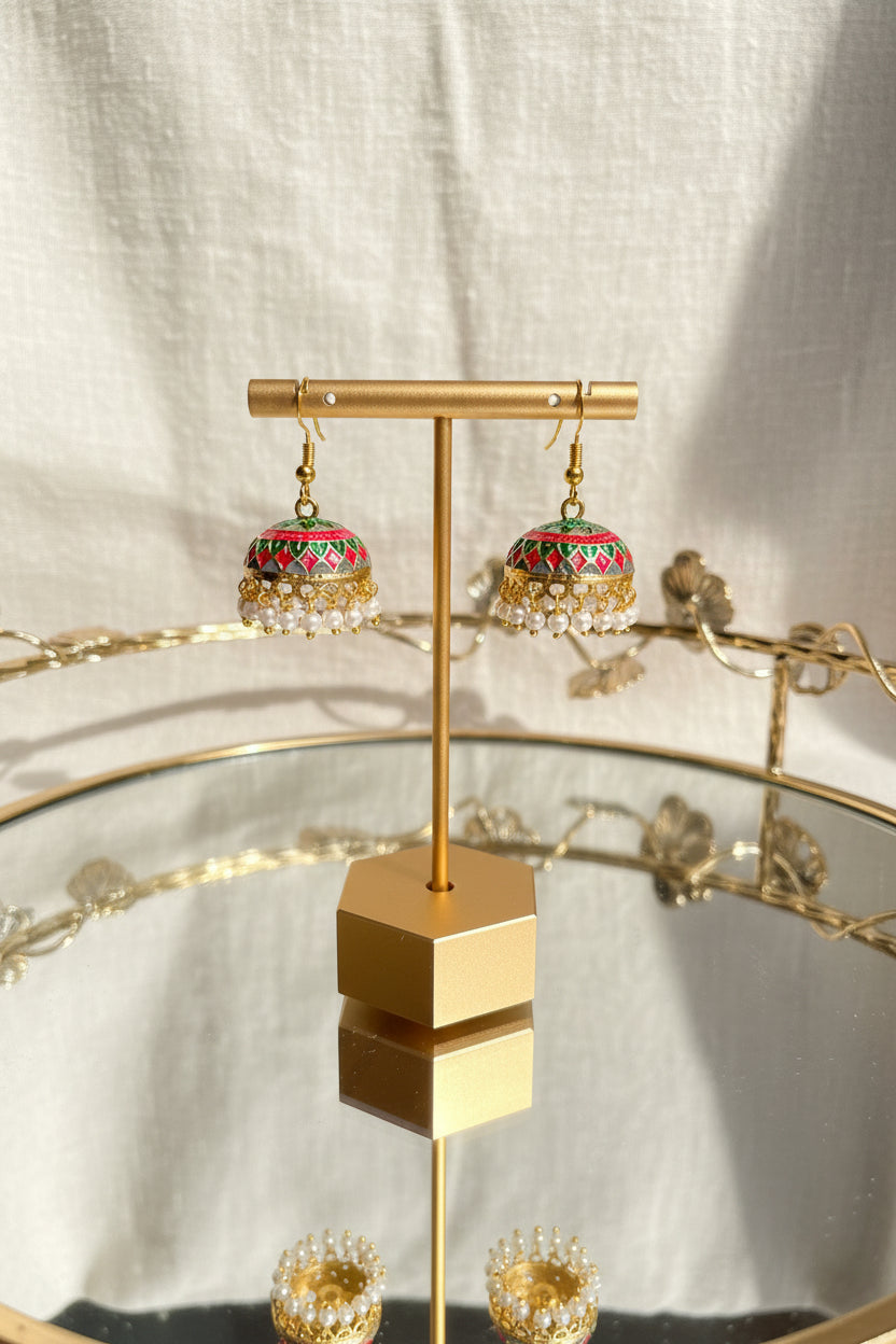 A pair of ornate gold-toned Jaipuri jhumkha earrings with red, green, and blue enamel detailing and white bead fringe, displayed on a gold stand over a reflective glass table.