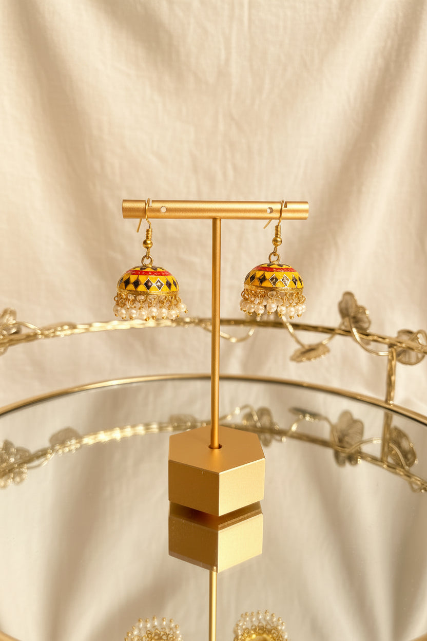 A pair of gold-toned Jaipuri jhumkha earrings with colorful enamel detailing and white pearl fringe, displayed on a gold stand against a white fabric backdrop.