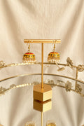 A pair of gold-toned Jaipuri jhumkha earrings with colorful enamel detailing and white pearl fringe, displayed on a gold stand against a white fabric backdrop.