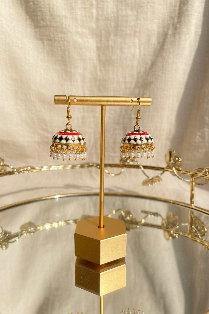 A pair of gold-toned Jaipuri jhumkha earrings with red, black, and white patterned tops and dangling pearl-like beads, displayed on a gold stand against a draped beige fabric background.