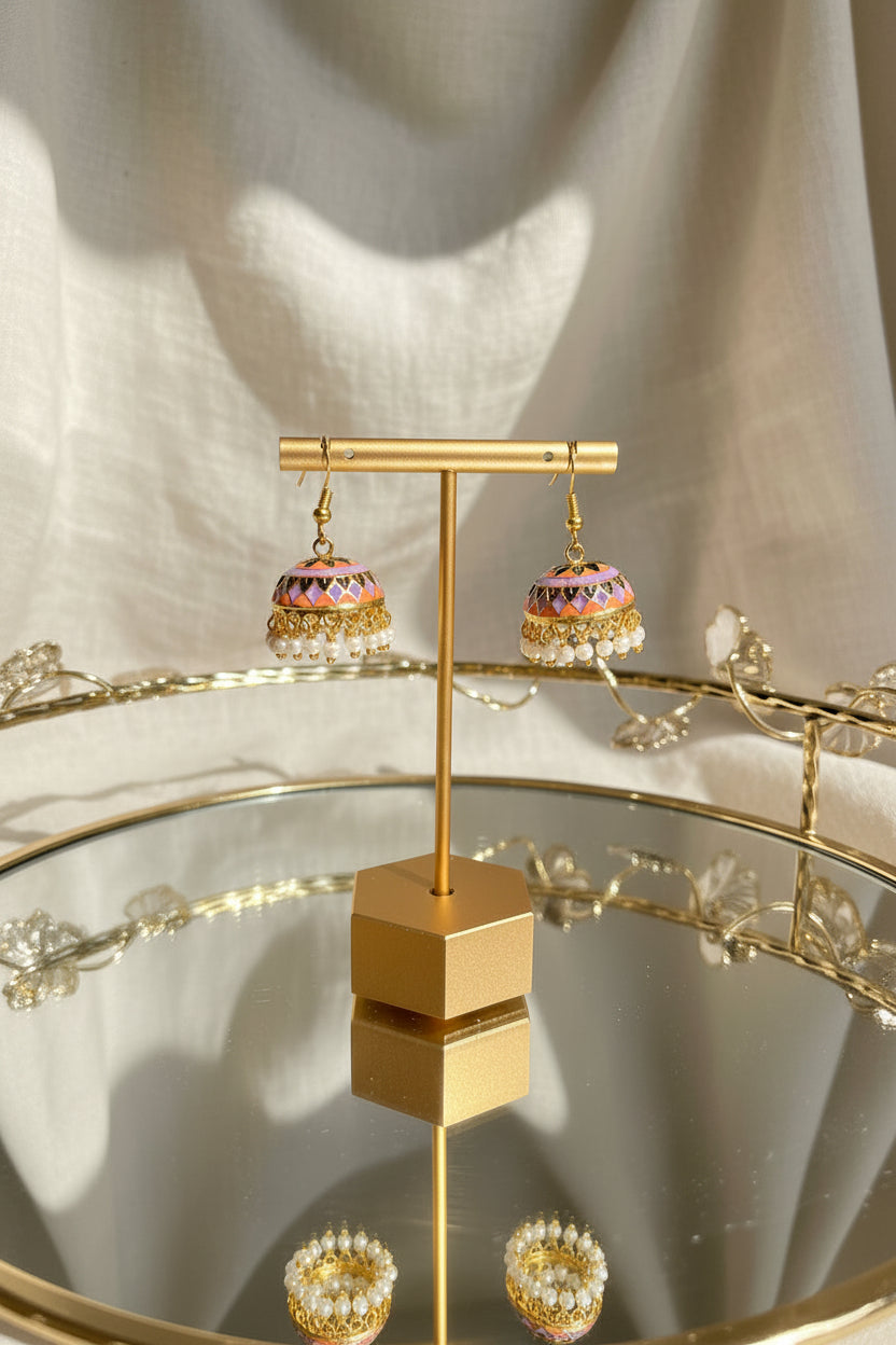 A pair of ornate gold-toned Jaipuri jhumkha earrings with colorful enamel detailing and white bead fringe, displayed on a gold stand on a reflective glass table with a window and greenery in the background.