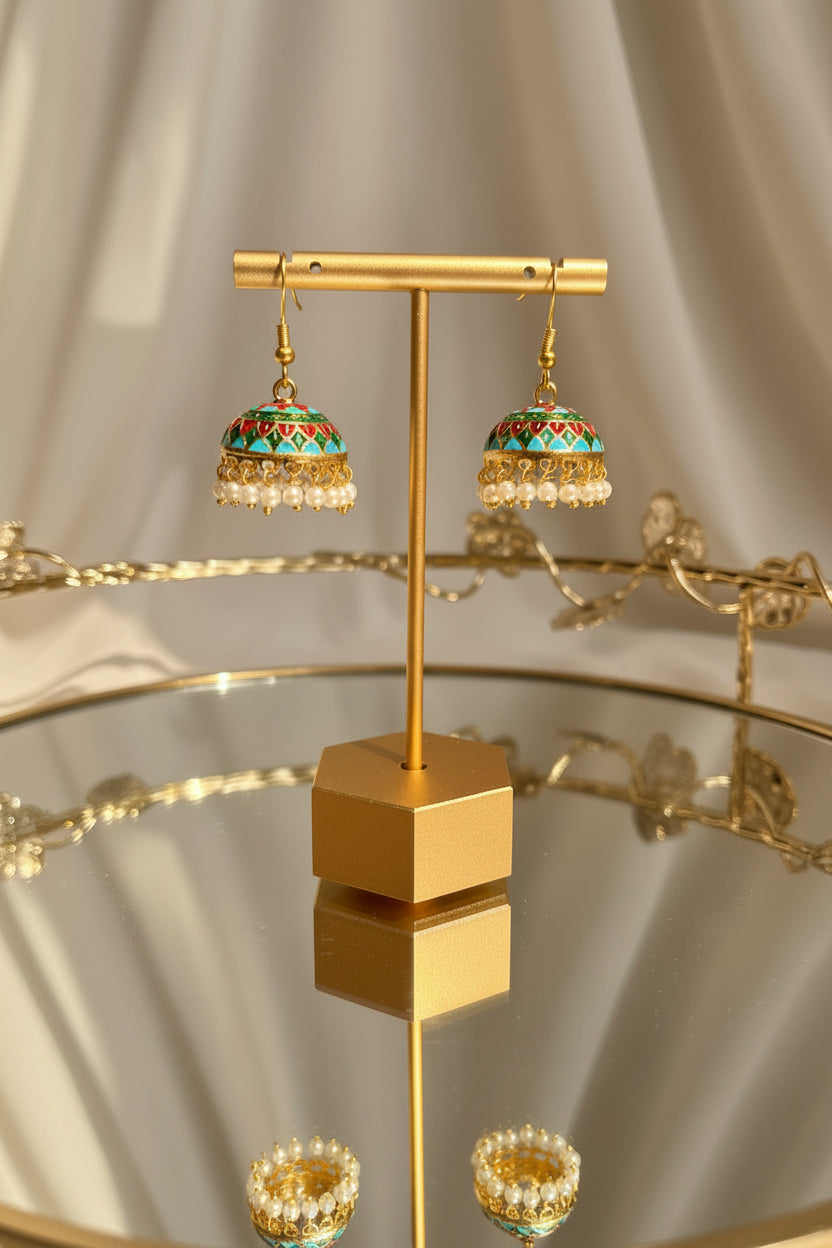A pair of ornate gold-toned Jaipuri jhumkha earrings with green, red, and white enamel detailing and pearl-like bead drops, displayed on a gold stand over a mirrored surface.
