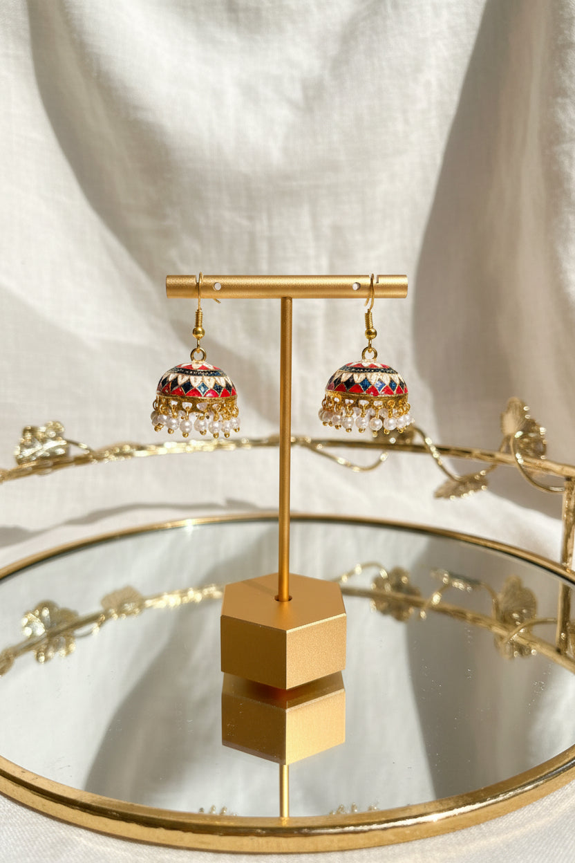 A pair of ornate gold-toned Jaipuri jhumkha earrings with colorful enamel detailing and white bead fringe, displayed on a gold stand on a mirrored tray.