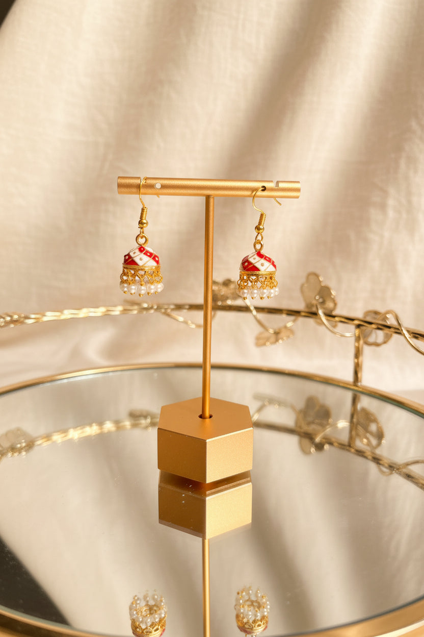 Gold-toned dangle Jaipuri jhumkha with red and white patterned fabric and small pearl-like beads, displayed on a gold stand over a mirrored tray against a soft beige fabric backdrop.
