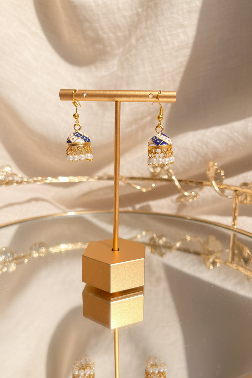 Gold-toned Jaipuri jhumkha earrings with blue and white enamel detailing and white bead drops, displayed on a gold stand on a reflective glass table.