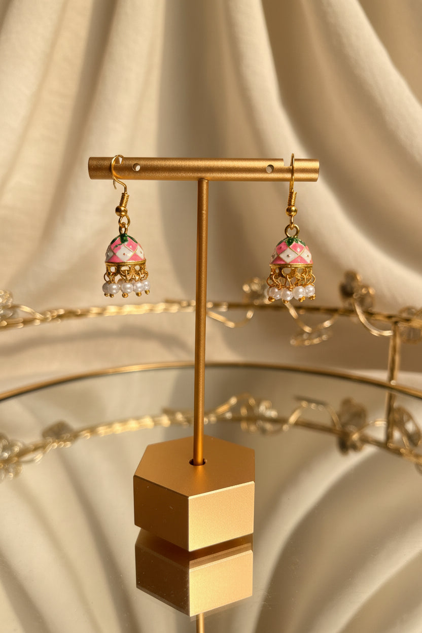 A pair of gold-toned dangle Jaipuri jhumkha with pink and white floral bell-shaped charms and small white bead drops, displayed on a gold stand on a reflective table with a blurred indoor background.
