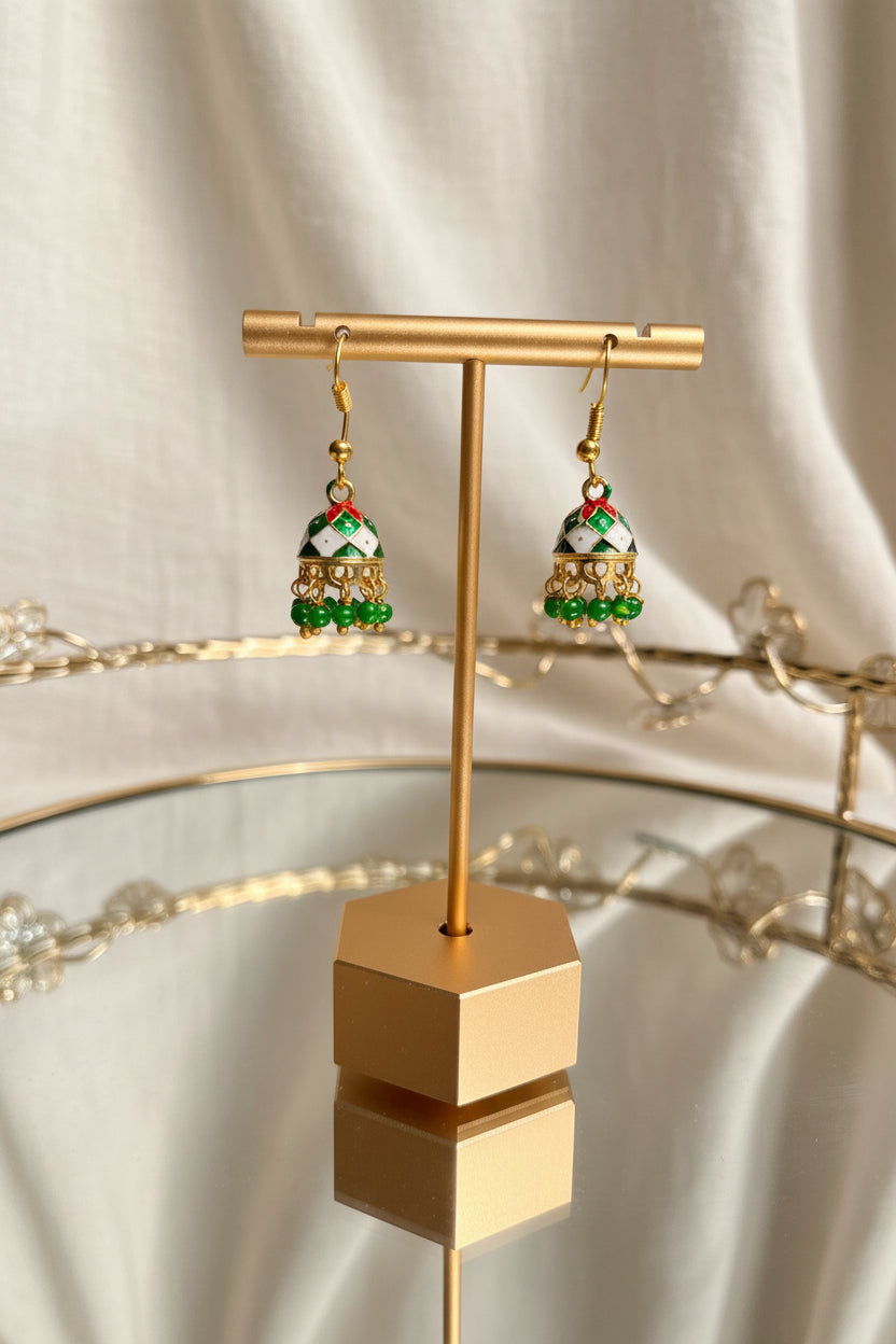 Gold-toned Jaipuri jhumkha earrings with white enamel, red accents, and green bead drops, displayed on a gold stand on a glass table.