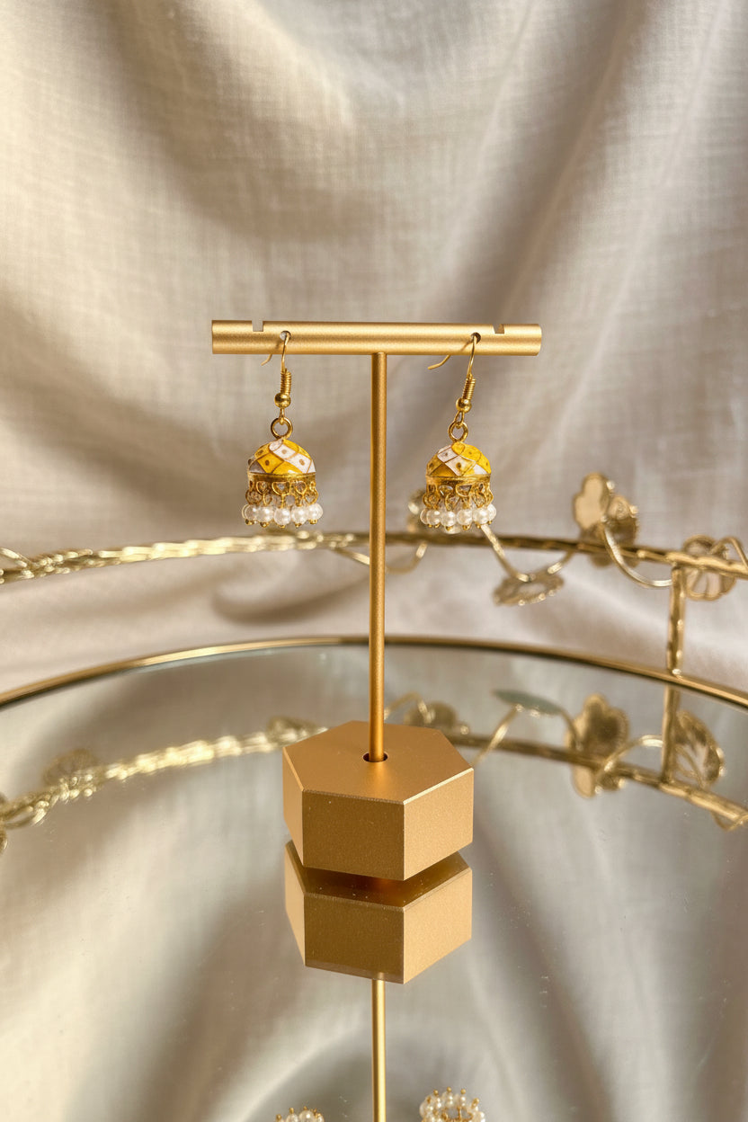A pair of ornate gold-toned jhumka Jaipuri jhumkha with white bead fringe and small floral details, displayed on a gold stand over a reflective glass table.
