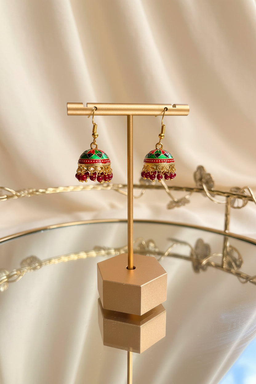 A pair of ornate gold-toned Jaipuri jhumkha earrings with red, green, and blue enamel detailing and dangling bead tassels, displayed on a gold stand on a glass table.