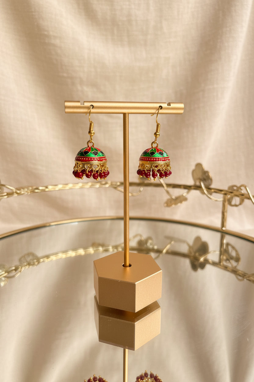 A pair of ornate gold-toned Jaipuri jhumkha earrings with red, green, and blue enamel detailing and dangling bead tassels, displayed on a gold stand on a glass table.