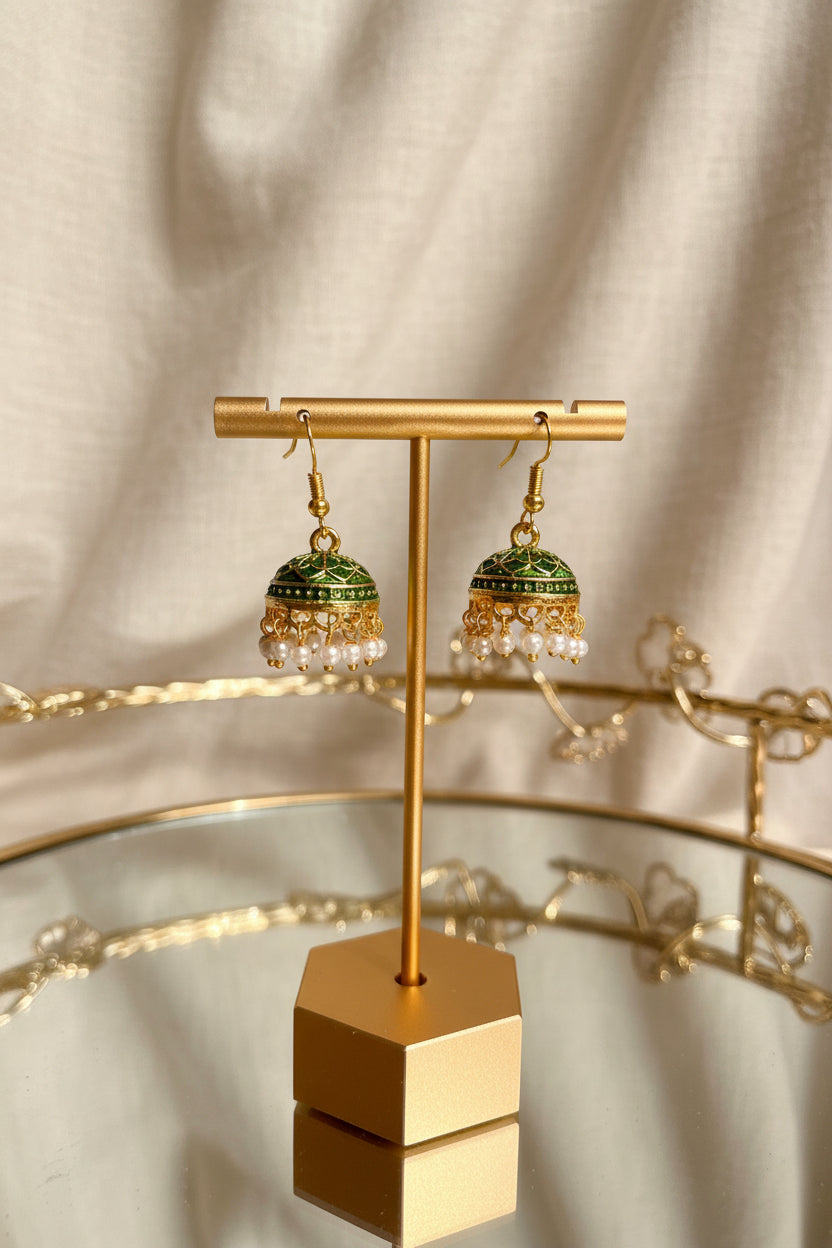 Gold-toned Jaipuri jhumkha earrings with green enamel detailing and white pearl drops, displayed on a gold stand against a soft beige fabric backdrop.