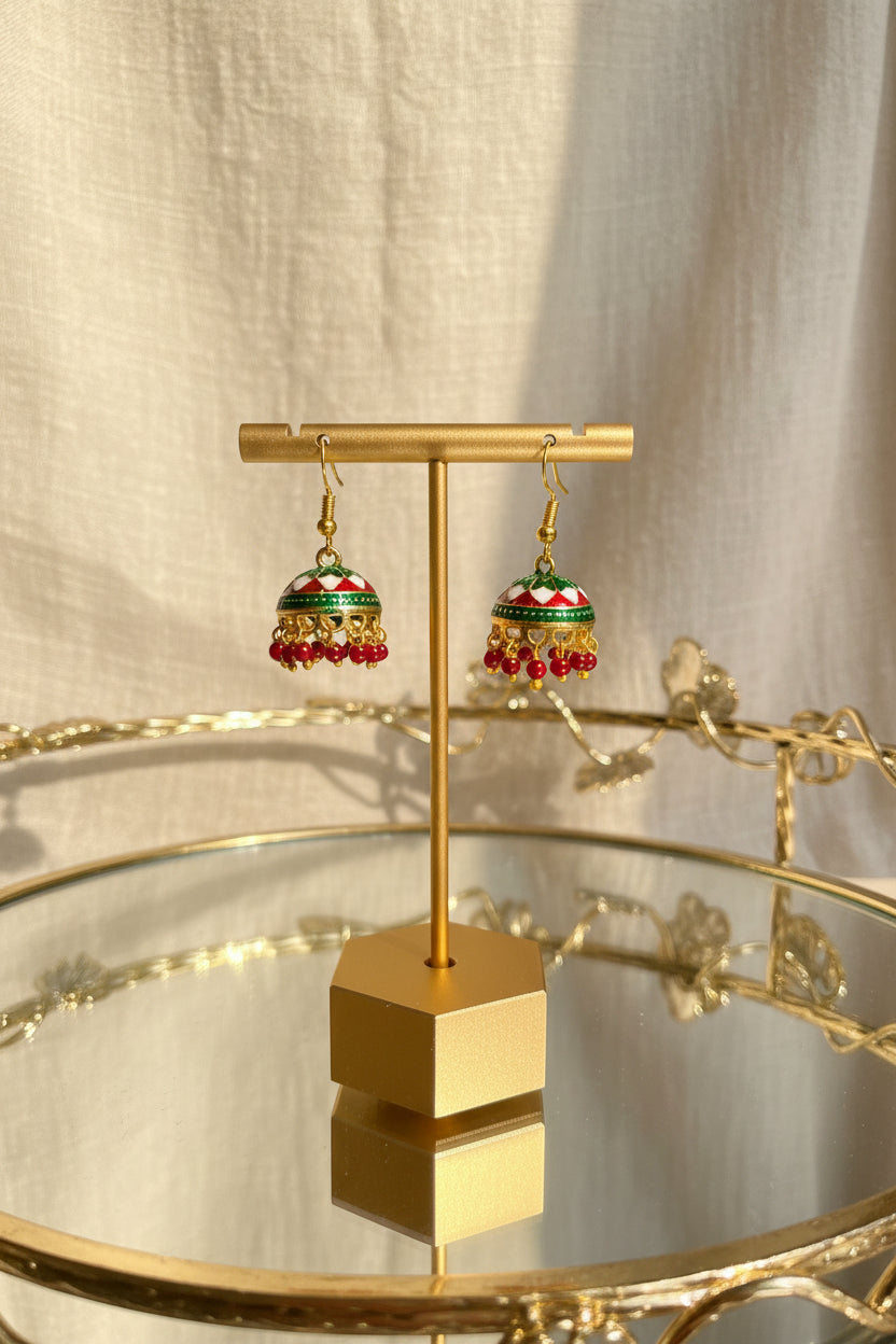 A pair of gold-toned Jaipuri jhumkha earrings with red, green, and white enamel detailing and dangling red beads, displayed on a gold stand over a mirrored tray against a cream fabric backdrop.