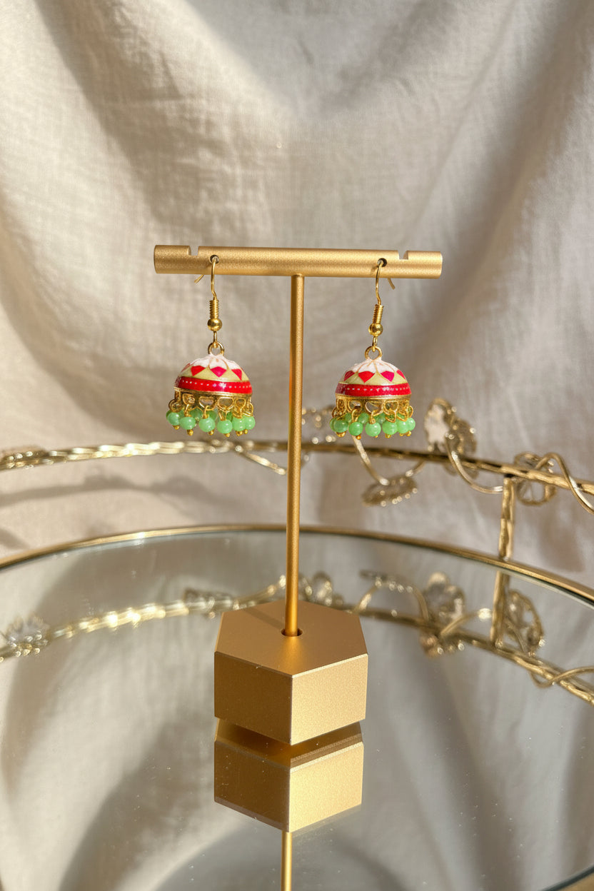 A pair of colorful traditional Indian Jaipuri jhumkha earrings displayed on a gold-colored stand. The earrings feature a red and pink patterned top, gold-toned metalwork, and dangling green bead tassels.
