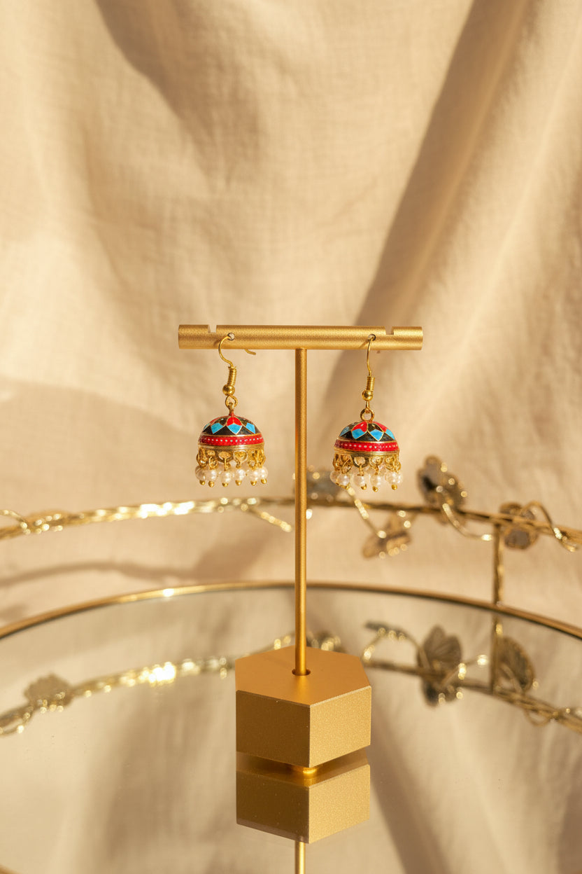 A pair of ornate gold-toned Jaipuri jhumkha earrings with colorful enamel detailing and dangling pearl-like beads, displayed on a gold stand against a draped beige fabric background.