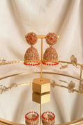 A pair of ornate gold-toned jhumka earrings with red stones and dangling red bead drops, displayed on a gold stand on a reflective table with a window and greenery in the background.