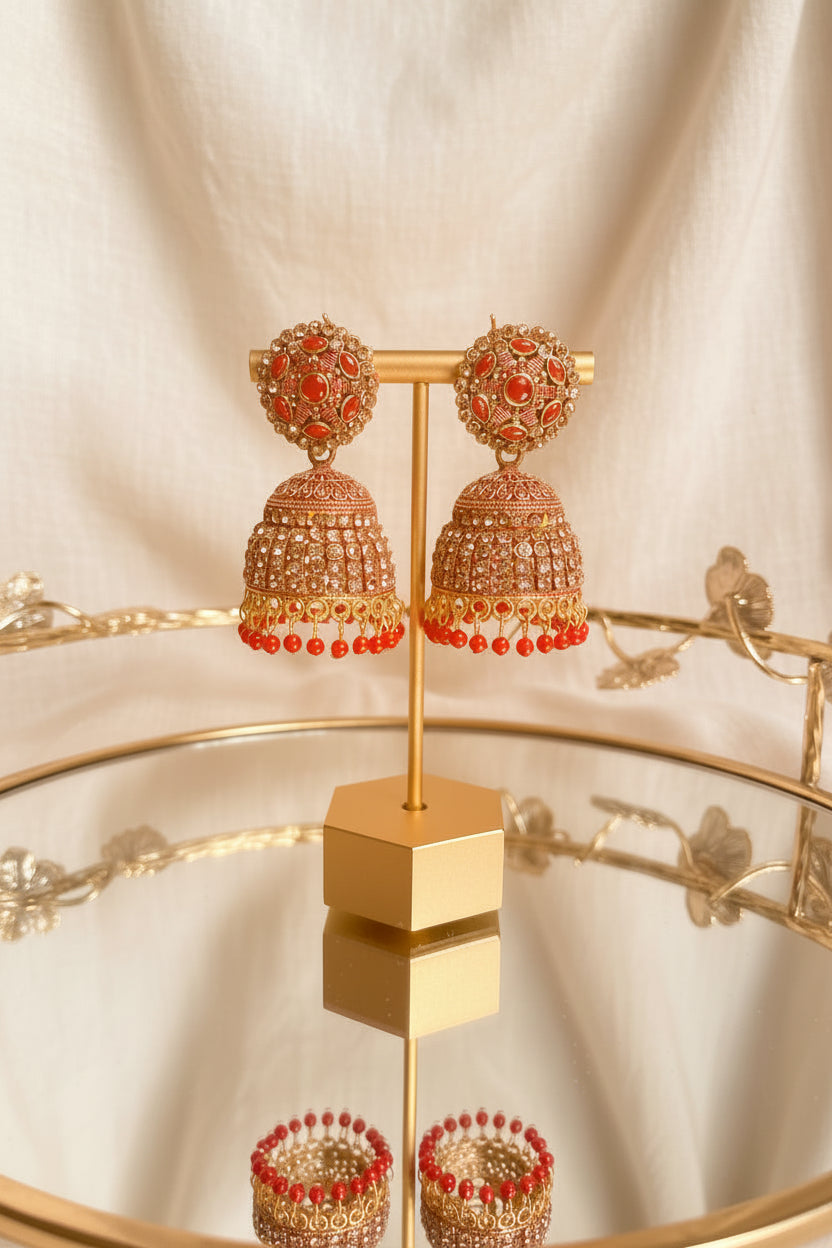 A pair of ornate gold-toned jhumka earrings with red stones and dangling red bead drops, displayed on a gold stand on a reflective table with a window and greenery in the background.