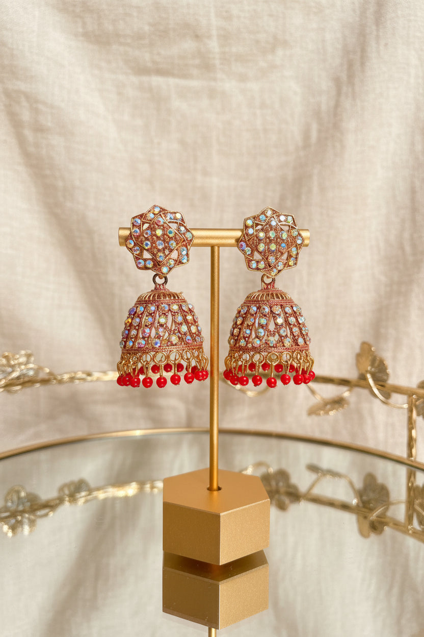 Gold-toned jhumka earrings with multicolored rhinestones and red dangling beads, displayed on a gold stand on a reflective glass table.