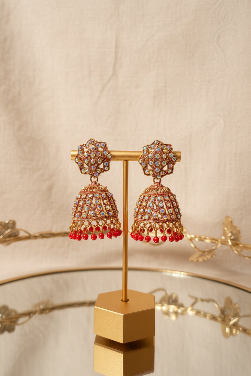 Gold-toned jhumka earrings with multicolored rhinestones and red dangling beads, displayed on a gold stand on a reflective glass table.