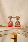 Gold-toned jhumka earrings with multicolored rhinestones and red dangling beads, displayed on a gold stand on a reflective glass table.