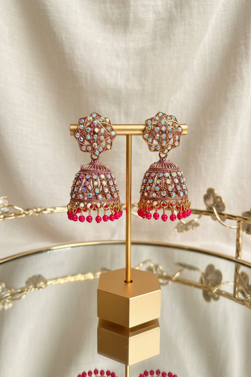A pair of ornate gold-toned jhumka earrings with pink and multicolor stone embellishments and dangling pink beads, displayed on a gold stand.