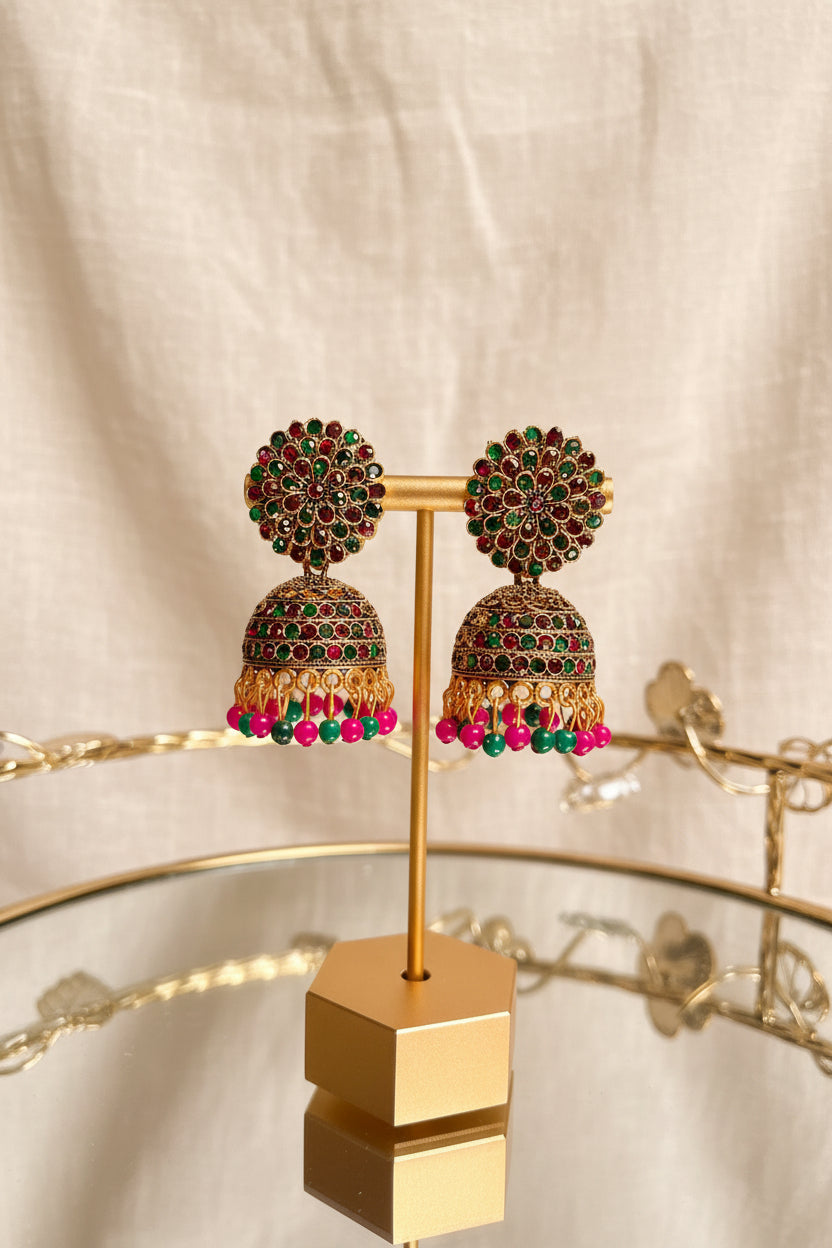 A pair of ornate gold-toned jhumka earrings with red and green stones and dangling pink and green beads, displayed on a gold stand on a glass table with a window and greenery in the background.