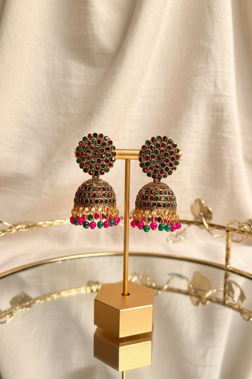 A pair of ornate gold-toned jhumka earrings with red and green stones and dangling pink and green beads, displayed on a gold stand on a glass table with a window and greenery in the background.