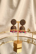 A pair of ornate gold-toned jhumka earrings with red and green stones and dangling pink and green beads, displayed on a gold stand on a glass table with a window and greenery in the background.