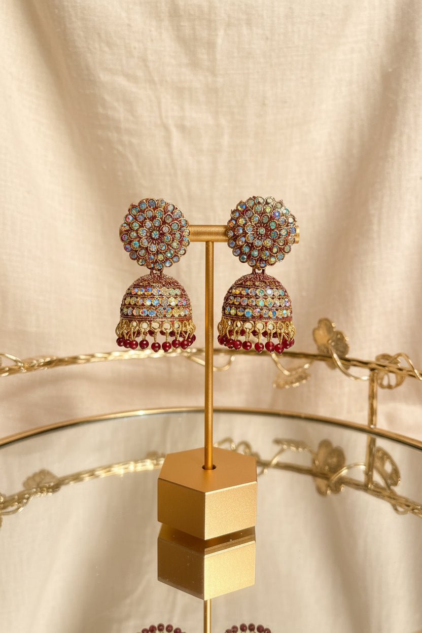 A pair of ornate gold-plated Indian jhumka earrings decorated with multicolored rhinestones, displayed on a gold stand on a glass table with a window background.