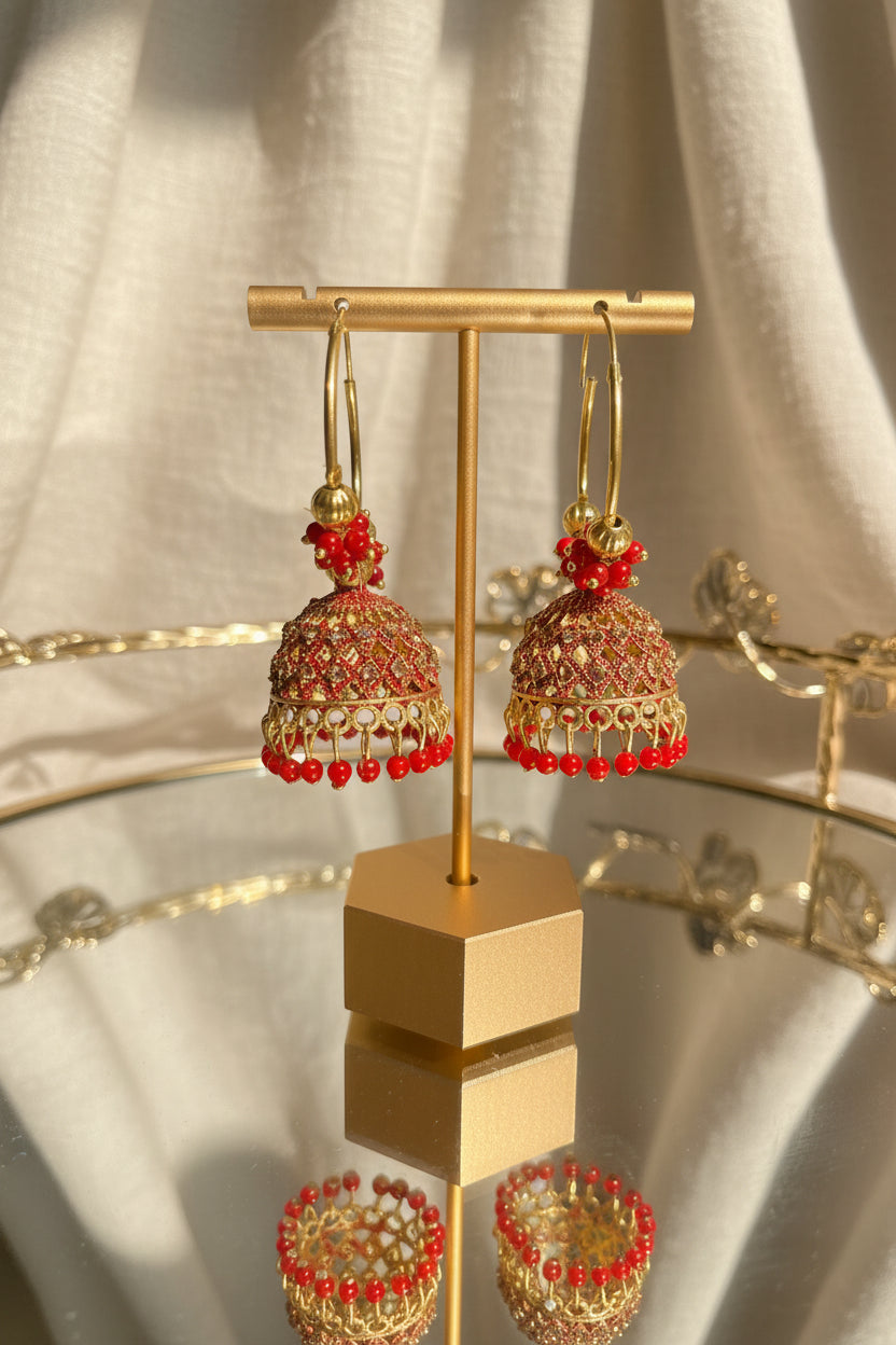 Gold and red beaded Indian hoop jhumka earrings hanging on a gold display stand on a glass table, with a blurred indoor background and greenery outside a window.