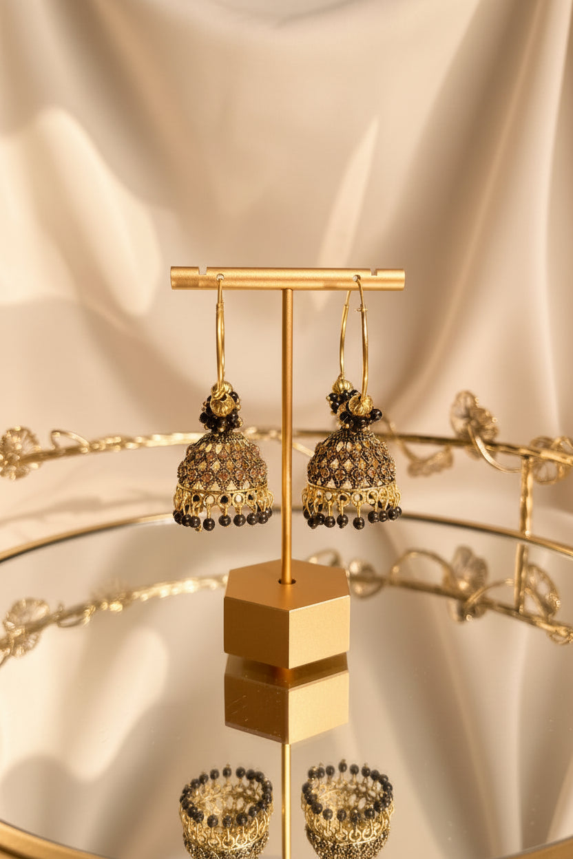 Gold-toned traditional hoop jhumka earrings with intricate beadwork and dangling beaded borders, displayed on a gold stand on a glass table with a blurred indoor background.