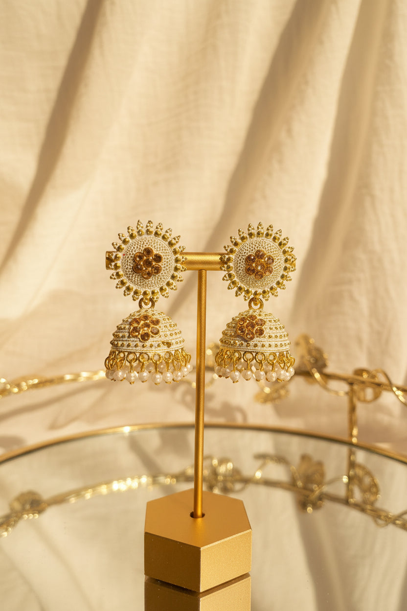 A pair of ornate gold and white jhumka earrings displayed on a gold stand, set against a softly blurred indoor background with a window and greenery.