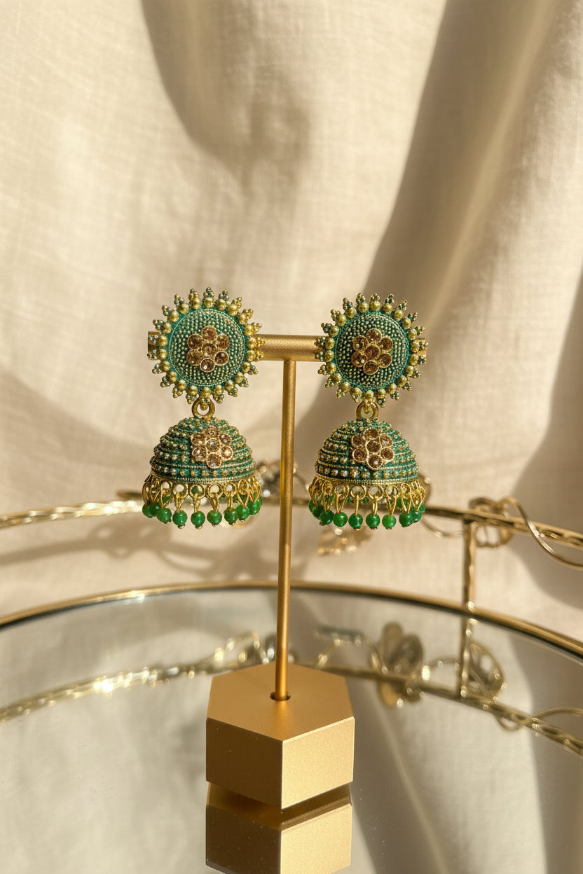 A pair of ornate Indian jhumka earrings with green enamel, gold-plated detailing, and small red stones, displayed on a gold stand on a glass table with a window in the background.