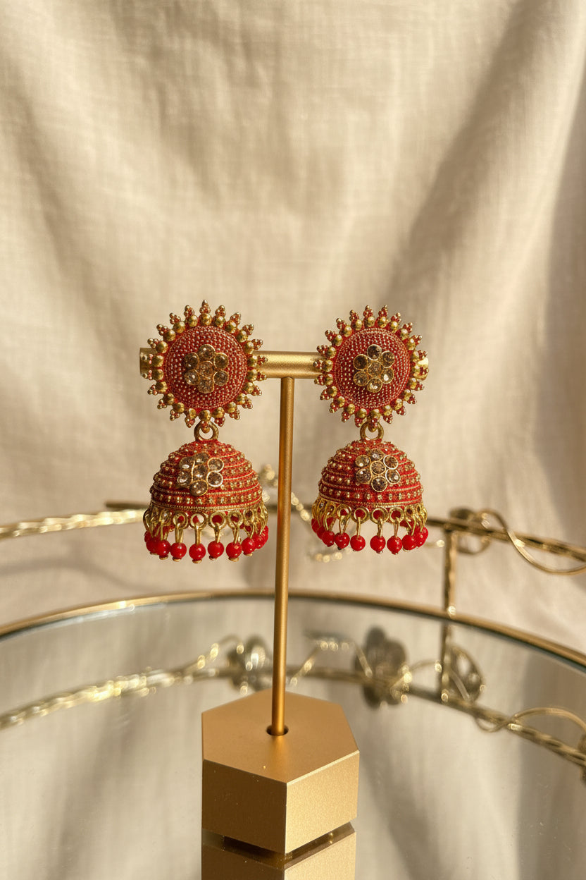 A pair of ornate red and gold jhumka earrings displayed on a gold stand, set against a softly blurred background of a window and greenery.