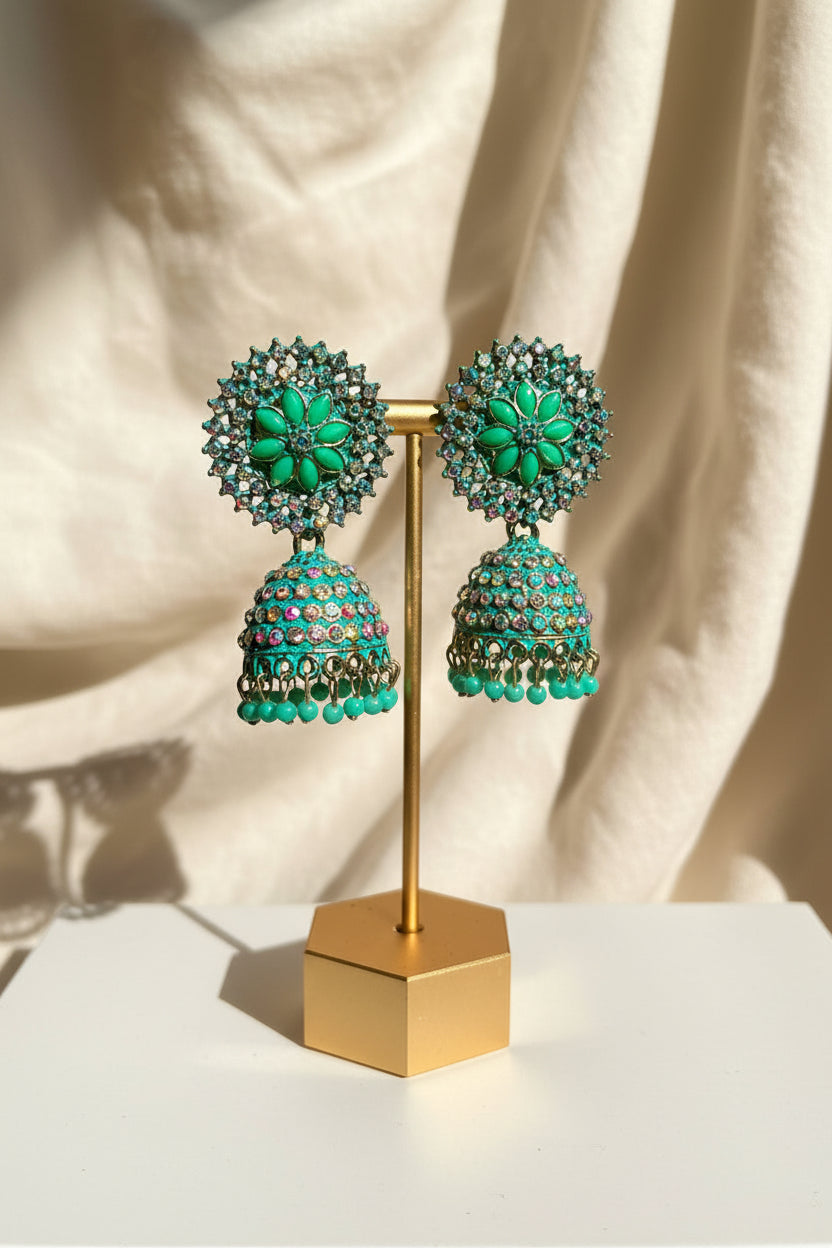 Turquoise and gold jhumka earrings displayed on a gold stand, held by a hand against a background of a beige wall and wooden trim.