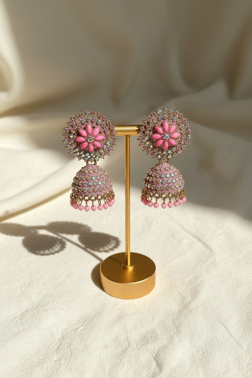 A pair of ornate pink and multicolor beaded jhumka earrings displayed on a gold stand, held by a hand against an indoor background with a wooden mantel and textured wall.