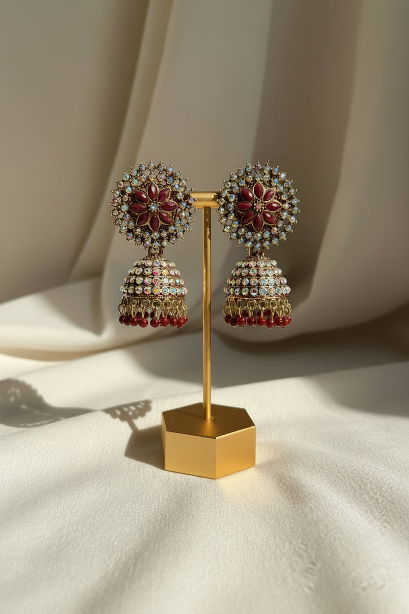 A pair of ornate Indian jhumka earrings displayed on a gold stand, held by a hand. The earrings feature gold-plated metal, deep red floral centers, and rows of iridescent blue and clear rhinestones, with dangling gold bells ending in red beads.