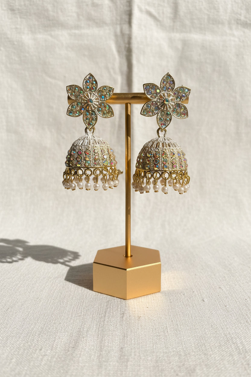 A pair of ornate gold-toned jhumka earrings with iridescent floral studs and dangling bead chains, displayed on a stand held by a hand against a blue fabric and wood background.
