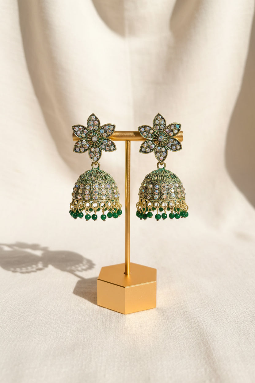 A pair of ornate gold-toned jhumka earrings with green and clear rhinestone embellishments, displayed on a gold stand held by a hand against an indoor background.