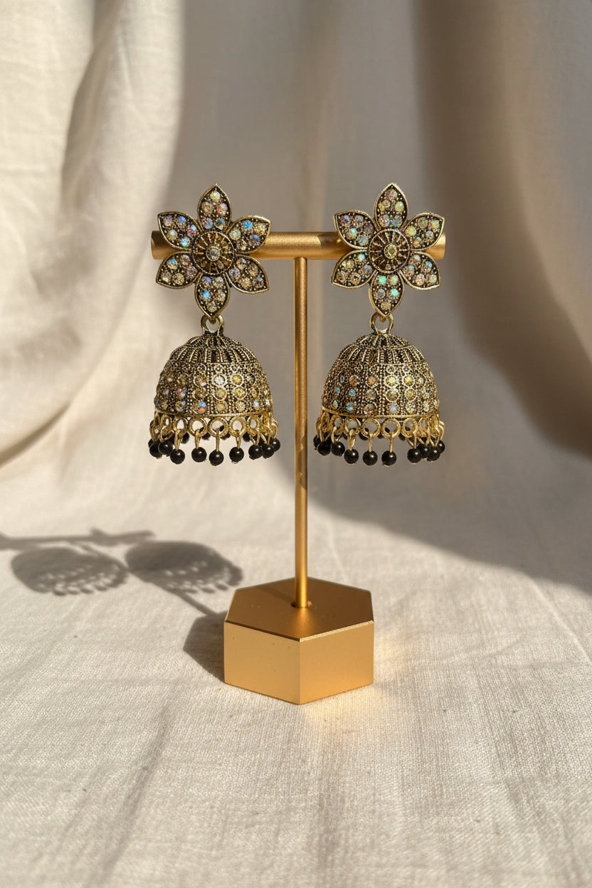 A pair of ornate gold-toned jhumka earrings with floral studs and bell-shaped dangling elements, displayed on a stand with a hand holding the base. The background shows a textured wall and blue fabric.