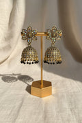 A pair of ornate gold-toned jhumka earrings with floral studs and bell-shaped dangling elements, displayed on a stand with a hand holding the base. The background shows a textured wall and blue fabric.