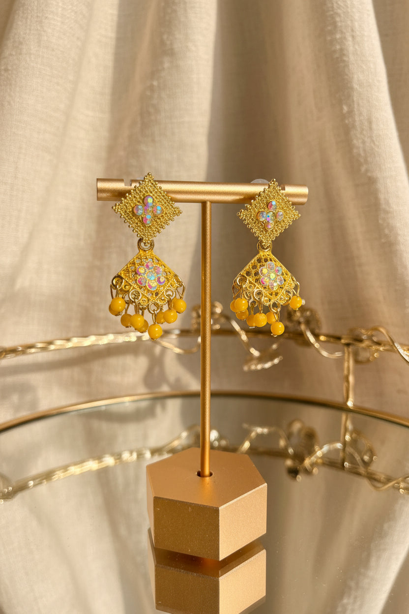 A pair of ornate gold-toned jhumka earrings with blue and yellow stones and dangling gold beads, displayed on a gold stand on a glass table with a blurred indoor background.