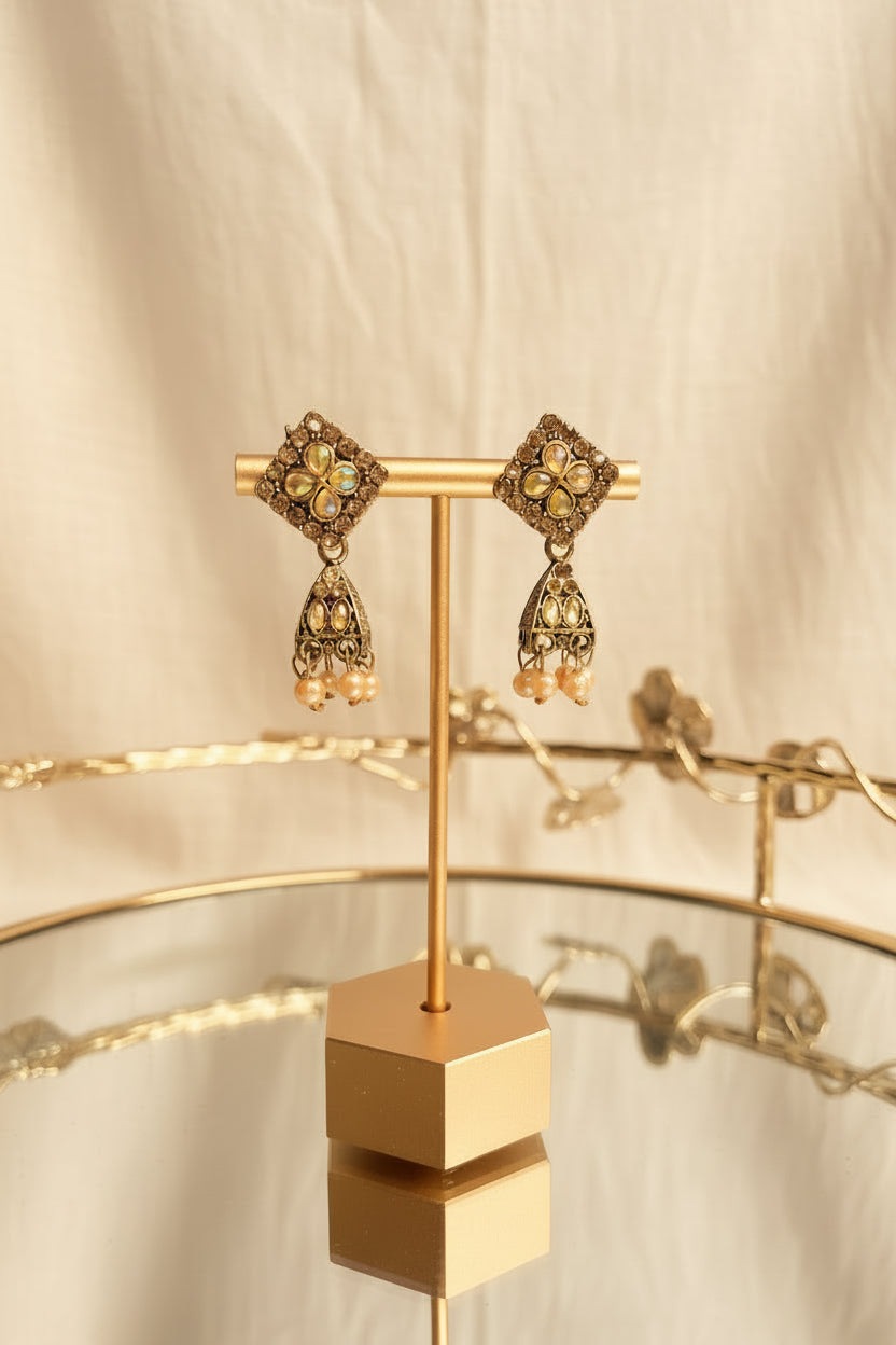 A pair of ornate gold-toned jhumka earrings displayed on a gold stand, featuring iridescent stones and small peach beads, with a blurred indoor background and window view.