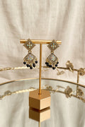 A pair of ornate gold-toned dangle earrings with light blue and white stones and black bead drops, displayed on a gold stand on a glass table with a blurred indoor background.
