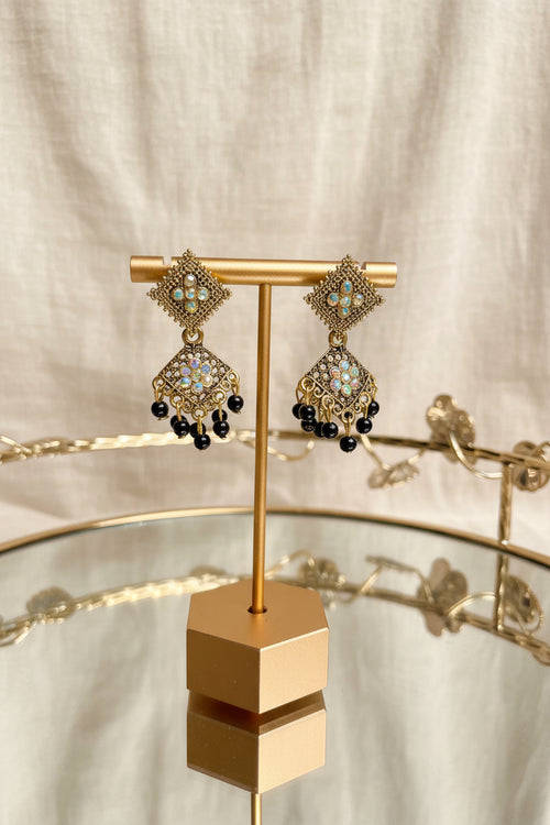 A pair of ornate gold-toned dangle earrings with light blue and white stones and black bead drops, displayed on a gold stand on a glass table with a blurred indoor background.