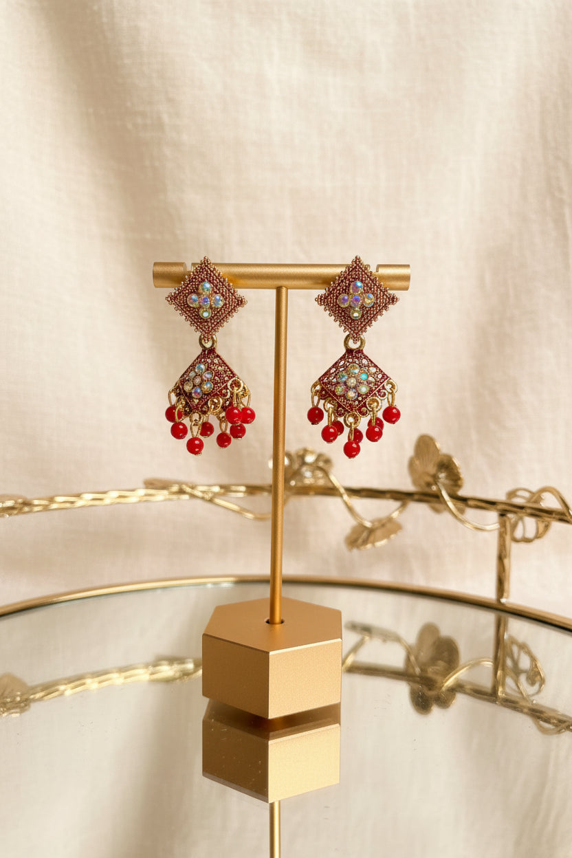A pair of ornate gold-toned chandbali earrings with red bead drops and light blue stone accents, displayed on a gold stand on a glass table with a window and greenery in the background.