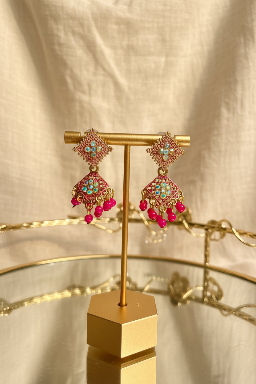 A pair of ornate gold-toned dangle earrings with pink and turquoise embellishments, displayed on a gold stand on a glass table with a blurred indoor background.